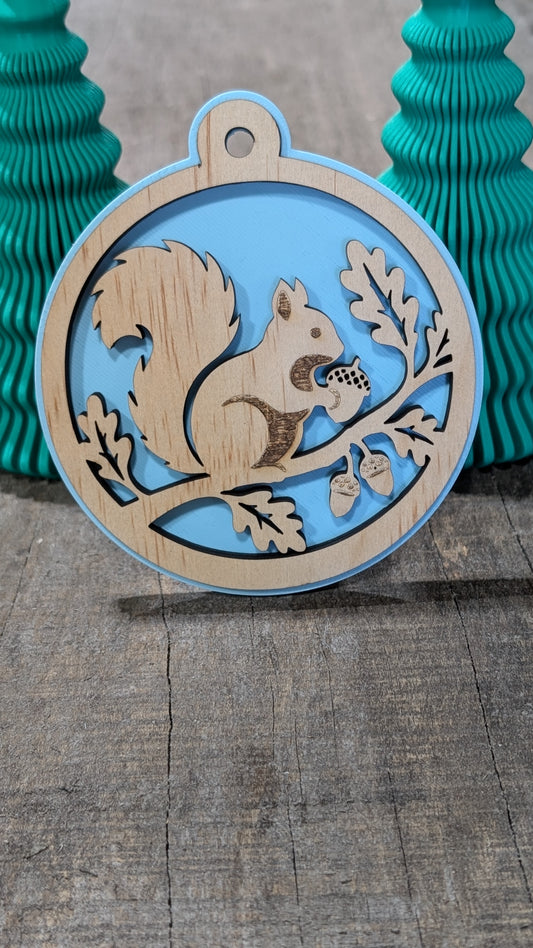 Boule de Noël en bois avec écureuil sur une branche avec glands et feuilles, sur fond bleu pâle.