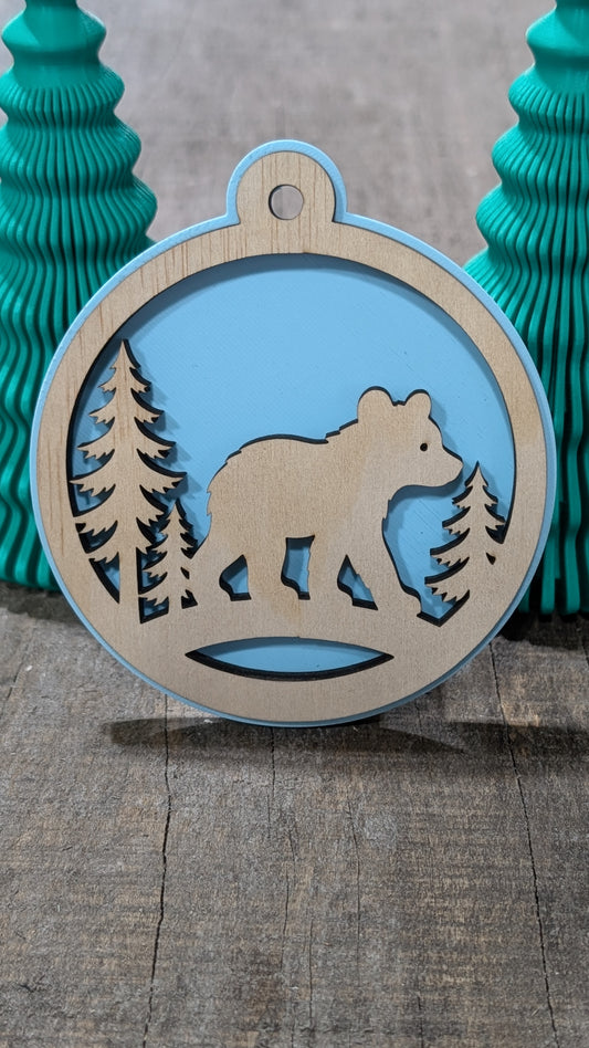 Boule de Noël avec ours et sapins en bois sur fond bleu pâle, fabriquée au Québec.
