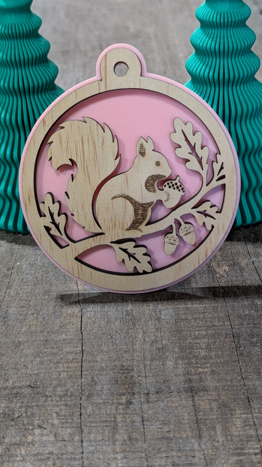 Boule de Noël en bois avec écureuil sur une branche avec glands et feuilles, sur fond rose.