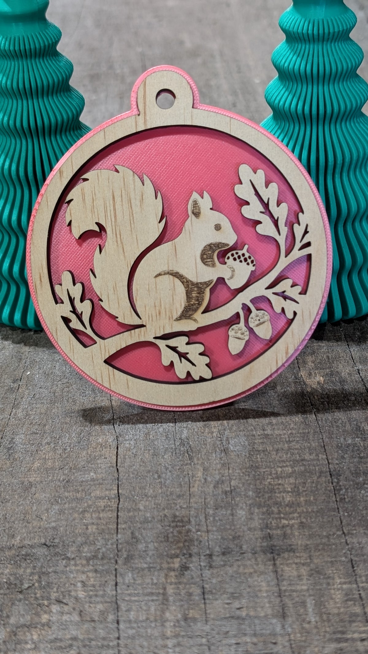 Boule de Noël en bois avec écureuil sur une branche avec glands et feuilles, sur fond rouge.