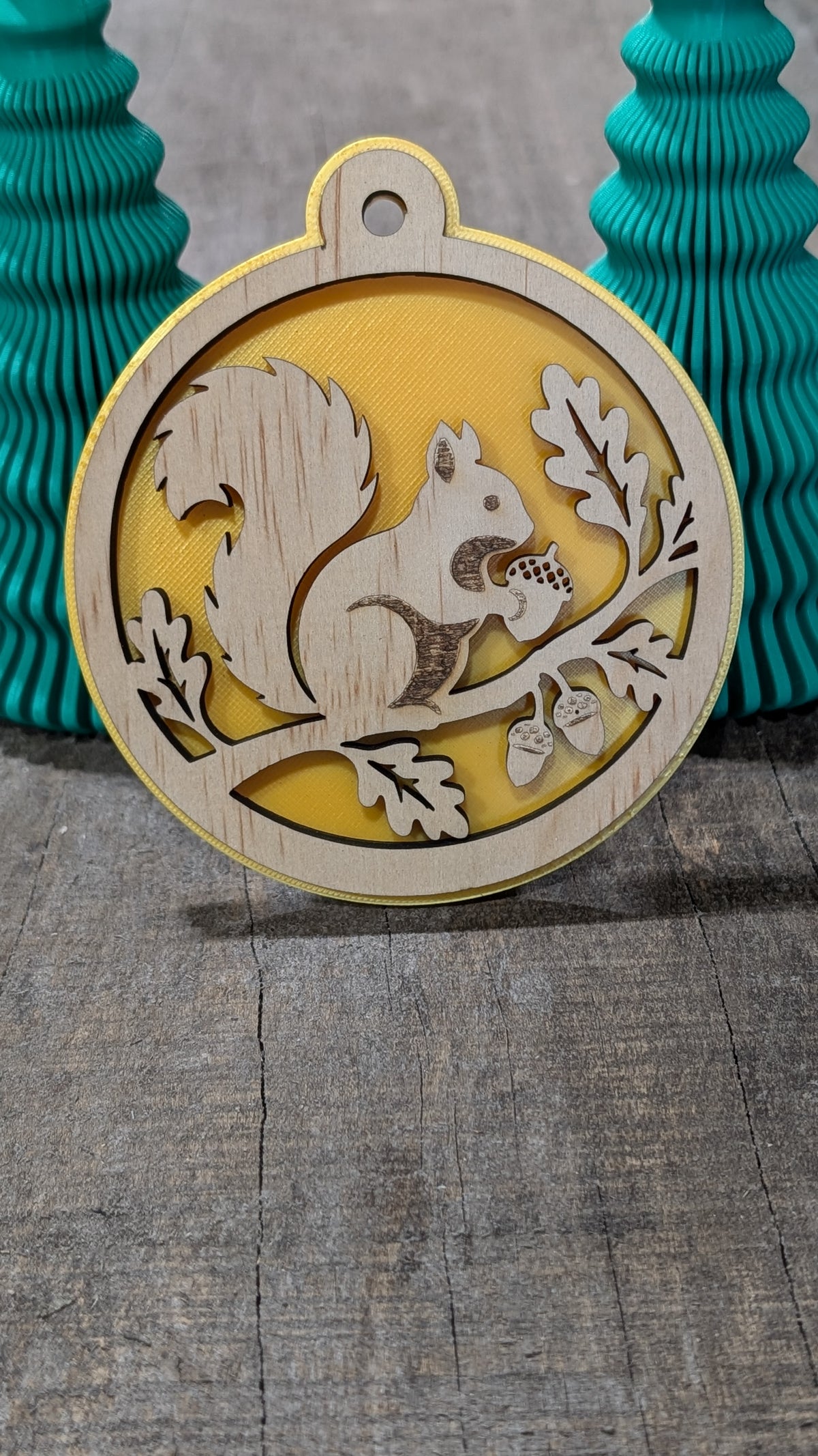 Boule de Noël en bois avec écureuil sur une branche avec glands et feuilles, sur fond jaune.