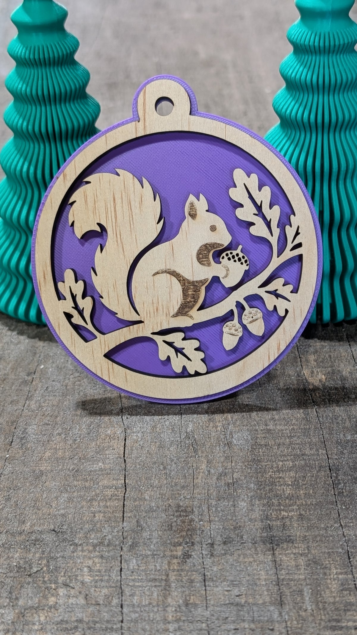 Boule de Noël en bois avec écureuil sur une branche avec glands et feuilles, sur fond mauve.