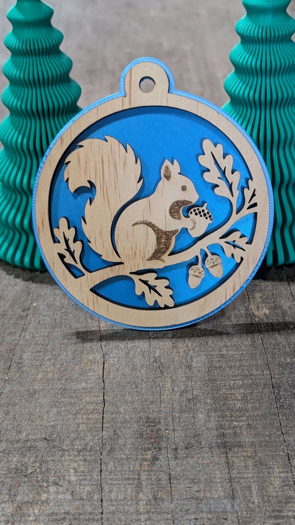 Boule de Noël en bois avec écureuil sur une branche avec glands et feuilles, sur fond bleu.
