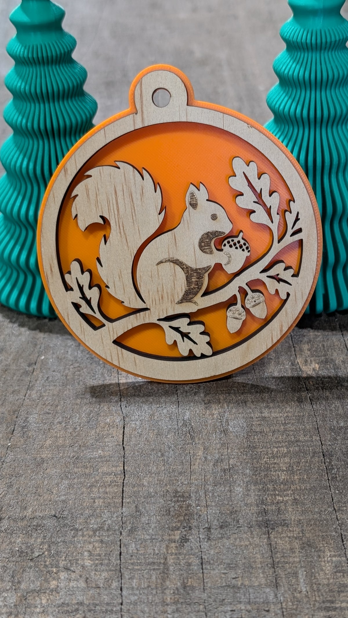 Boule de Noël en bois avec écureuil sur une branche avec glands et feuilles, sur fond orange.