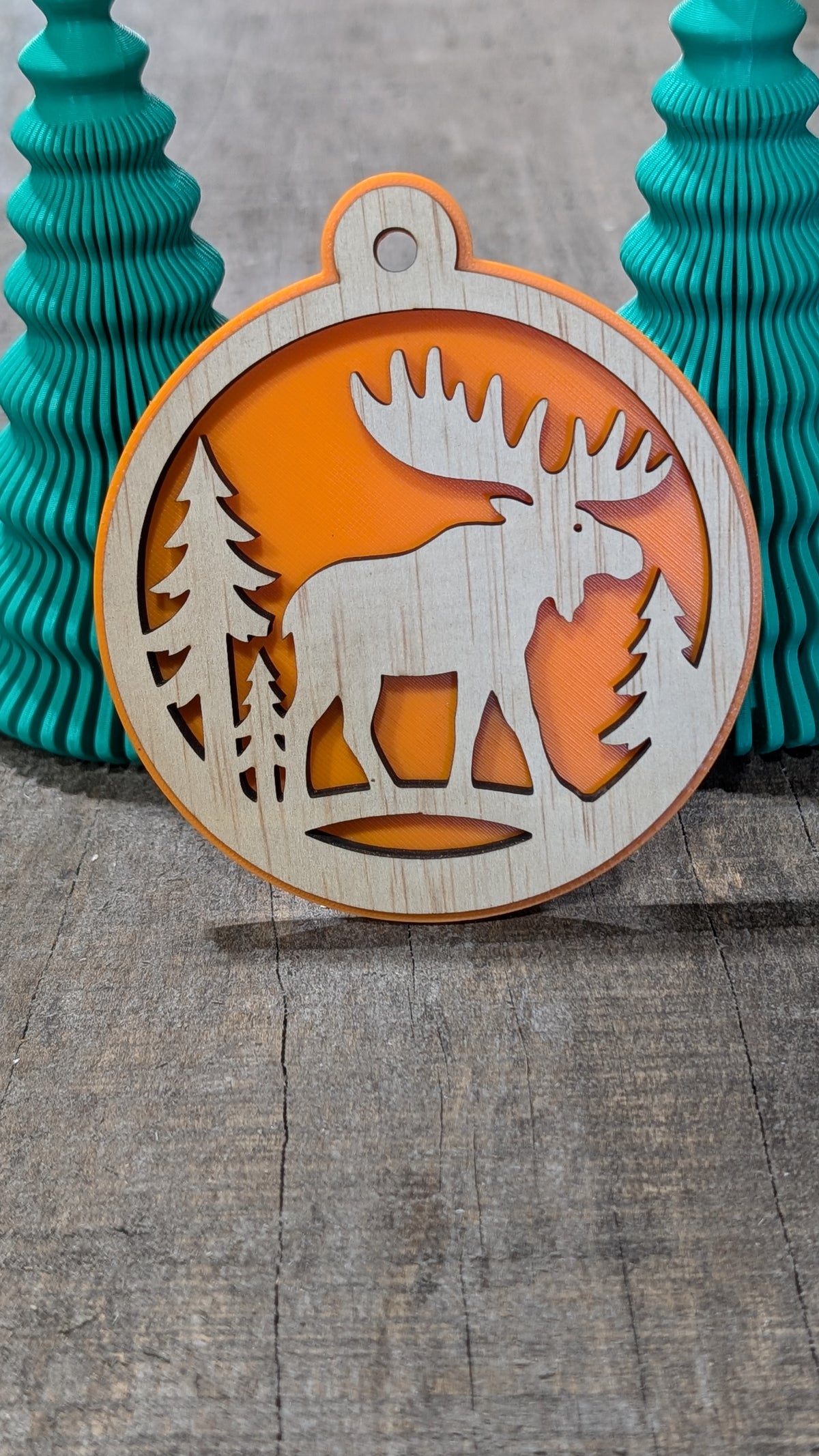 Boule de Noël en bois avec orignal et sapins, sur fond orange.