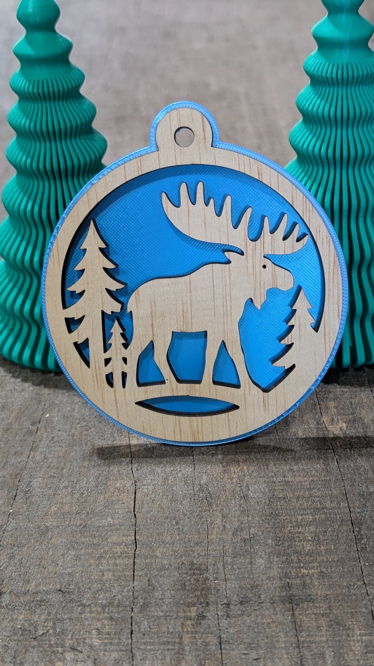 Boule de Noël en bois avec orignal et sapins, sur fond bleu.
