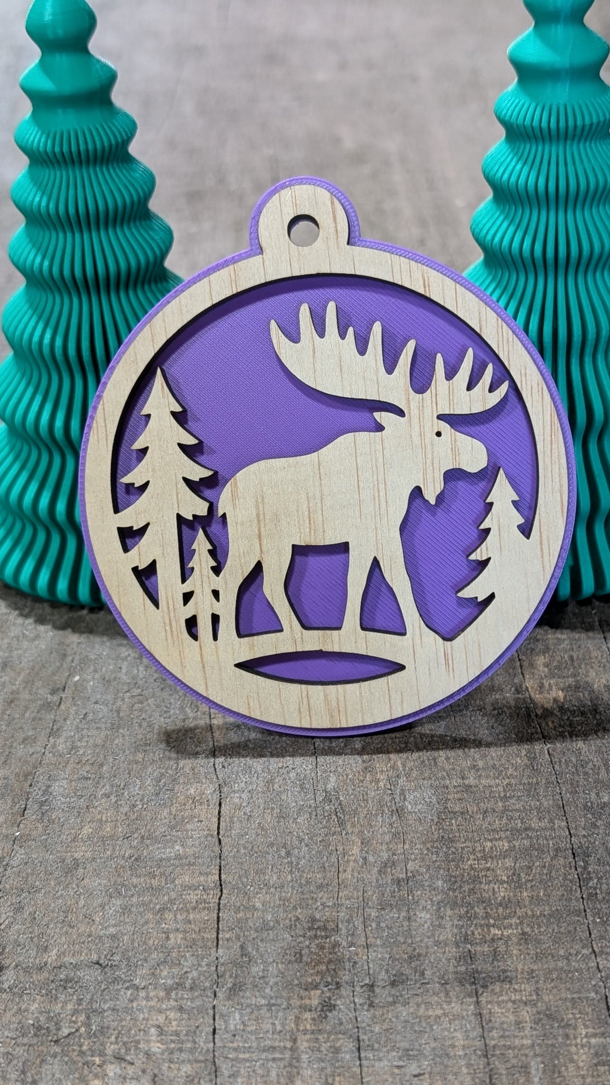 Boule de Noël en bois avec orignal et sapins, sur fond mauve.