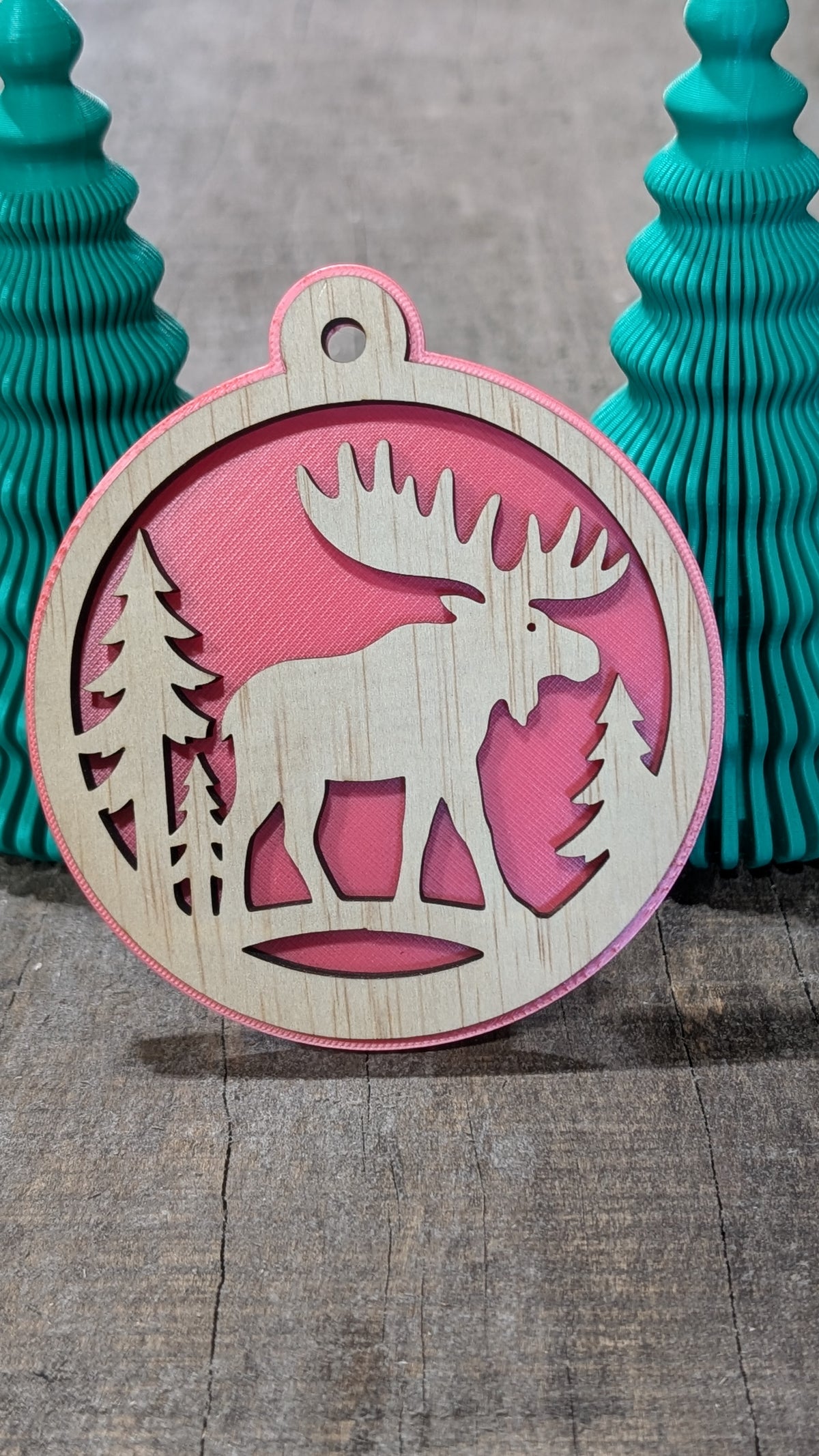 Boule de Noël en bois avec orignal et sapins, sur fond rouge.