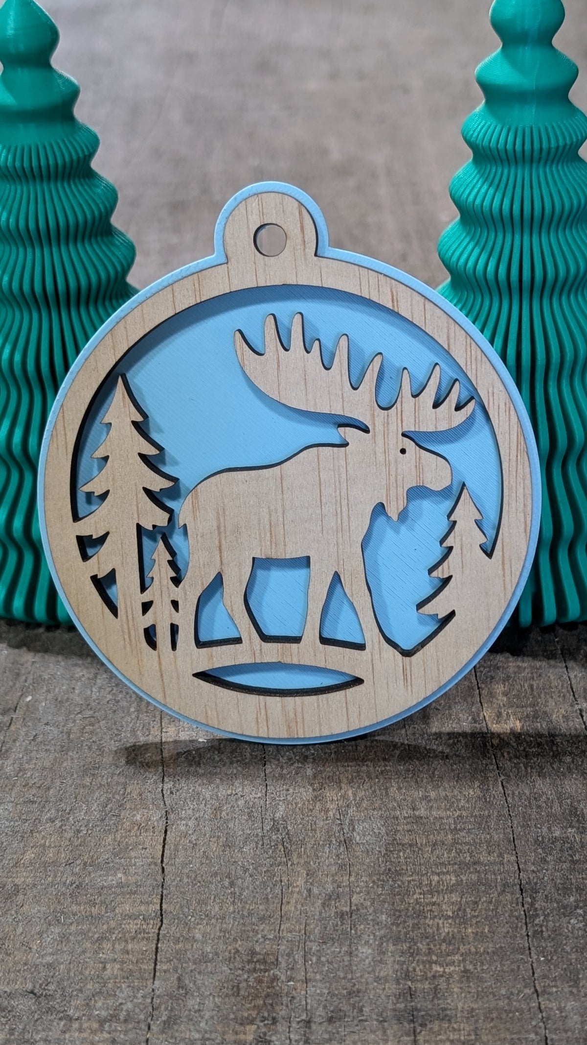 Boule de Noël en bois avec orignal et sapins, sur fond bleu pâle.