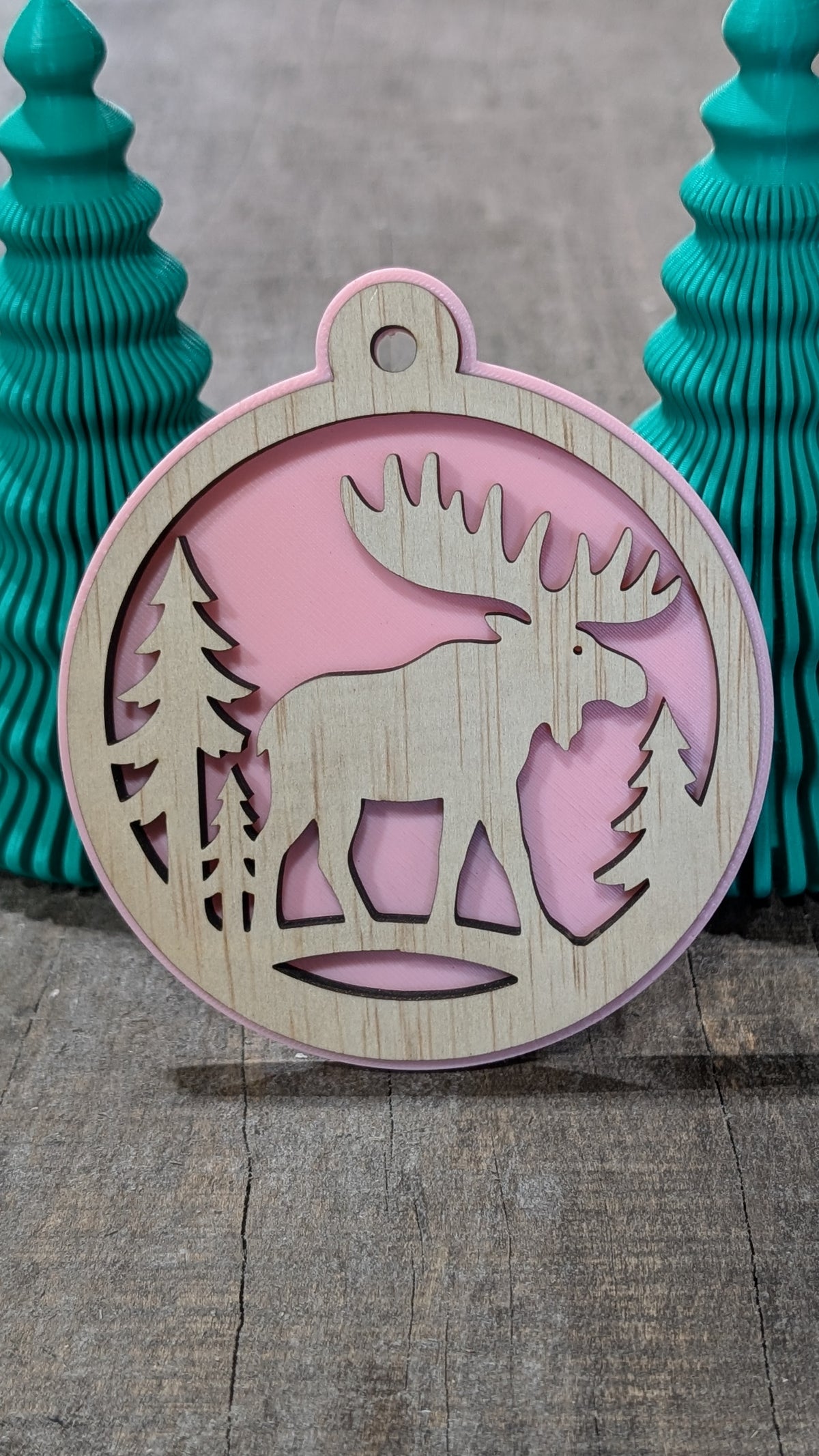 Boule de Noël en bois avec orignal et sapins, sur fond rose.