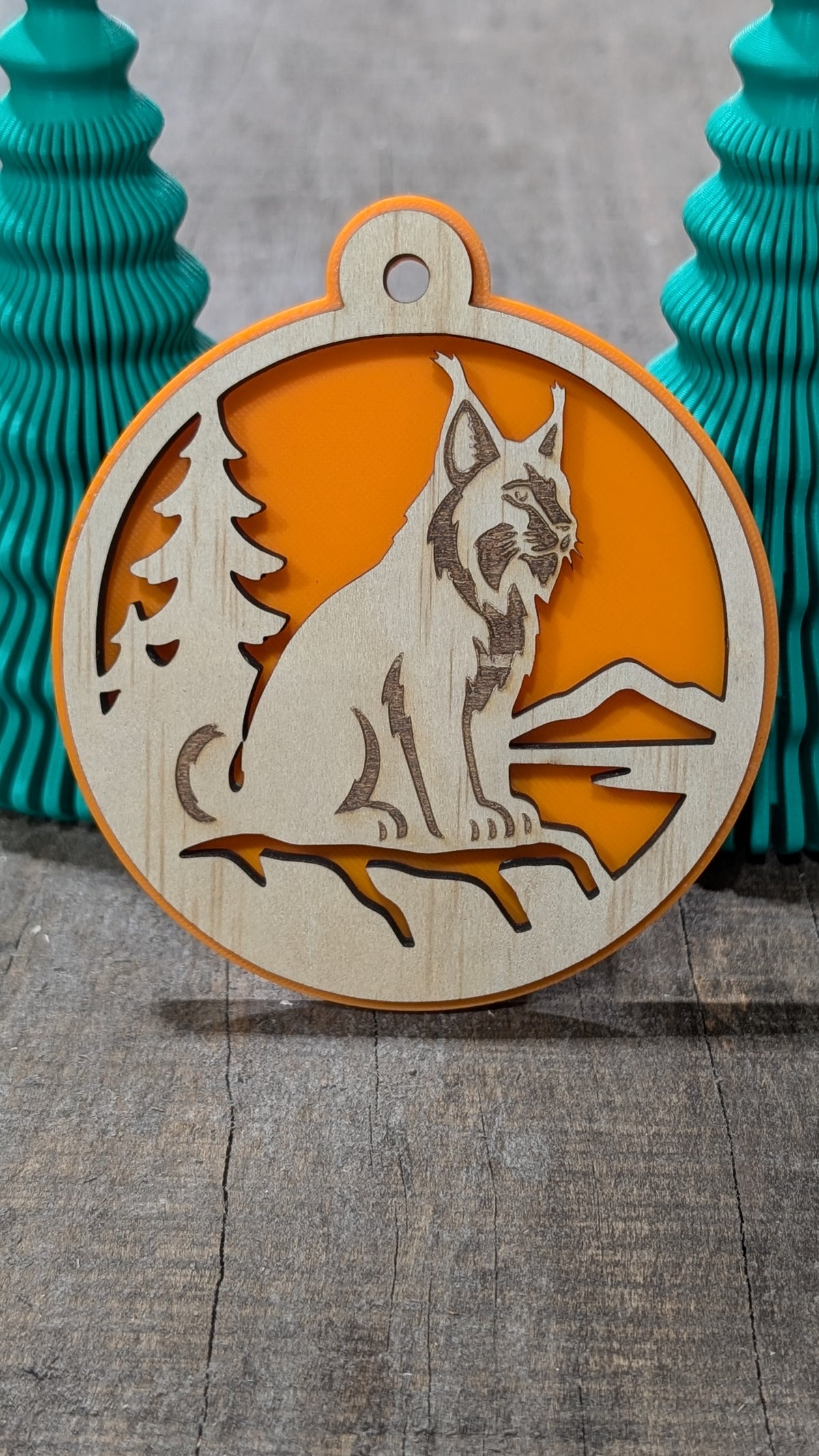 Boule de Noël en bois avec lynx, sapin et montagnes, sur fond orange.
