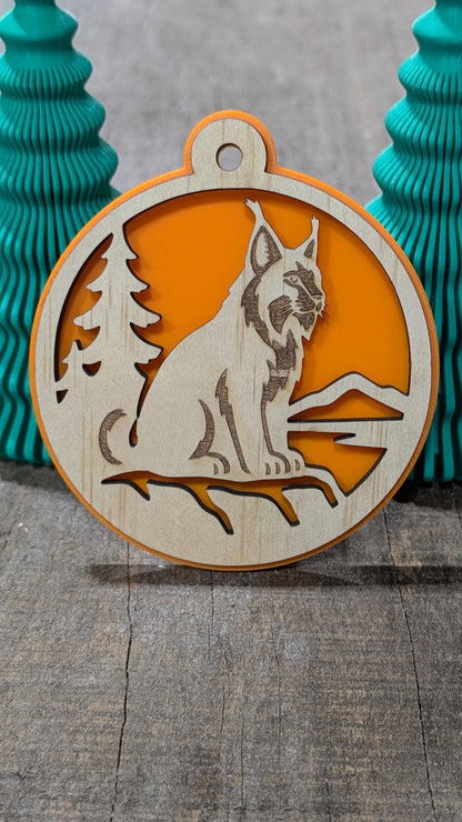 Boule de Noël en bois avec lynx, sapin et montagnes, sur fond orange.