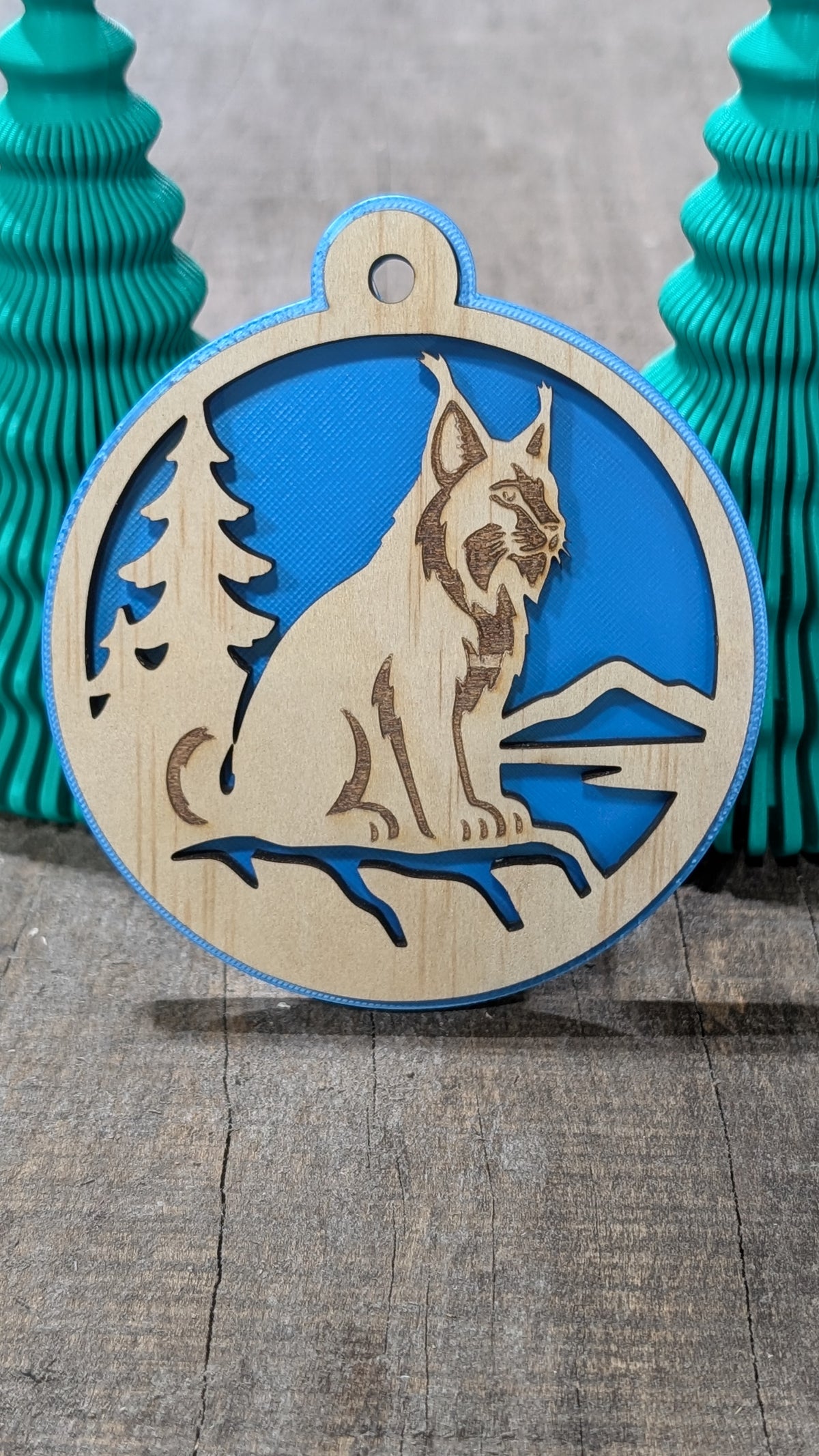 Boule de Noël en bois avec lynx, sapin et montagnes, sur fond bleu.