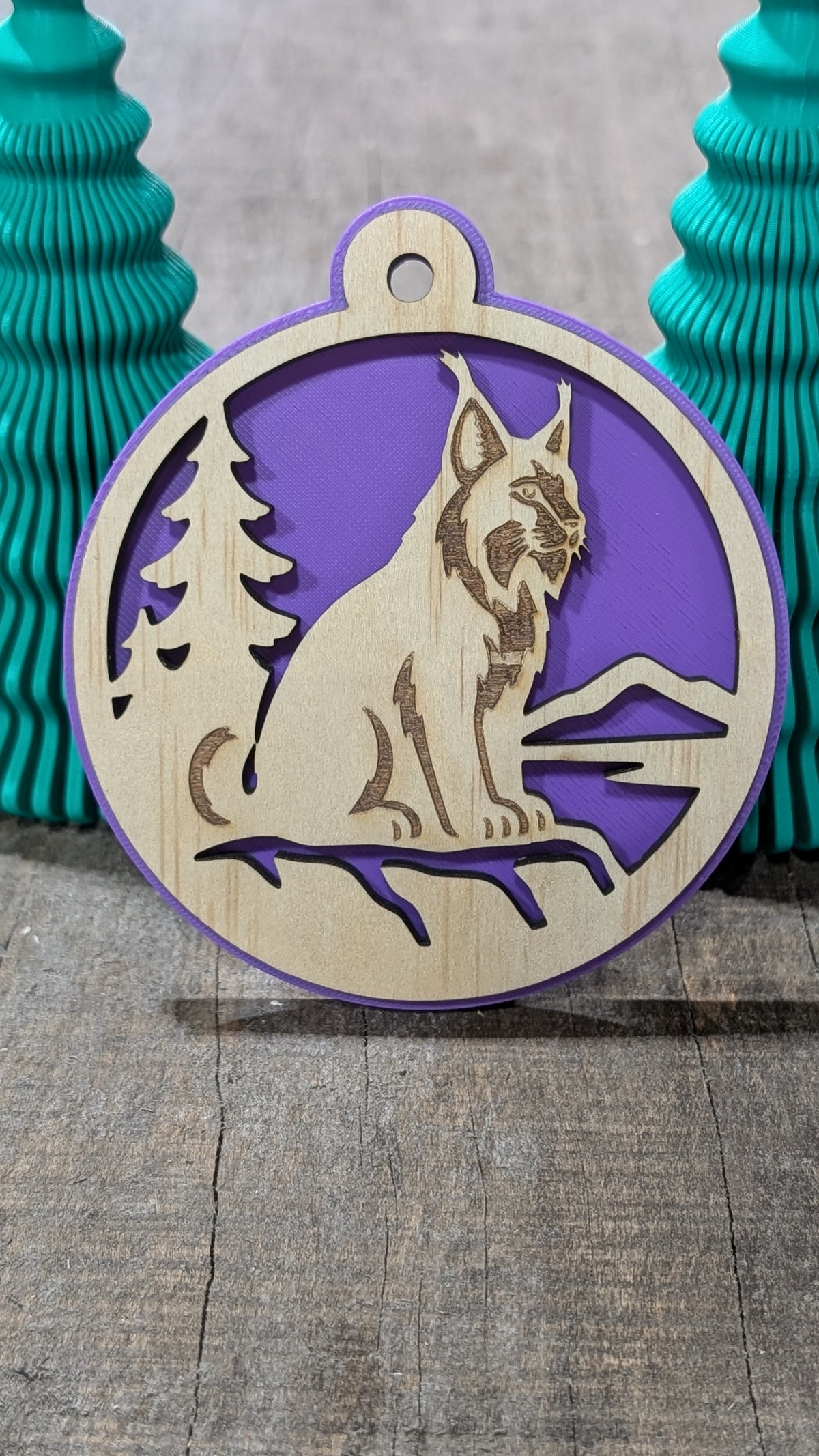 Boule de Noël en bois avec lynx, sapin et montagnes, sur fond mauve.