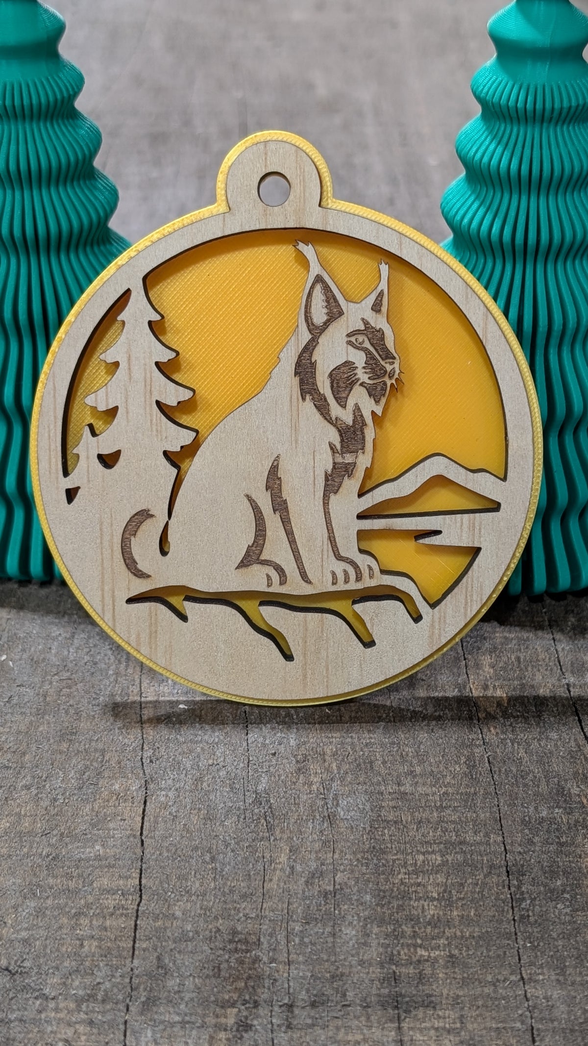 Boule de Noël en bois avec lynx, sapin et montagnes, sur fond jaune.