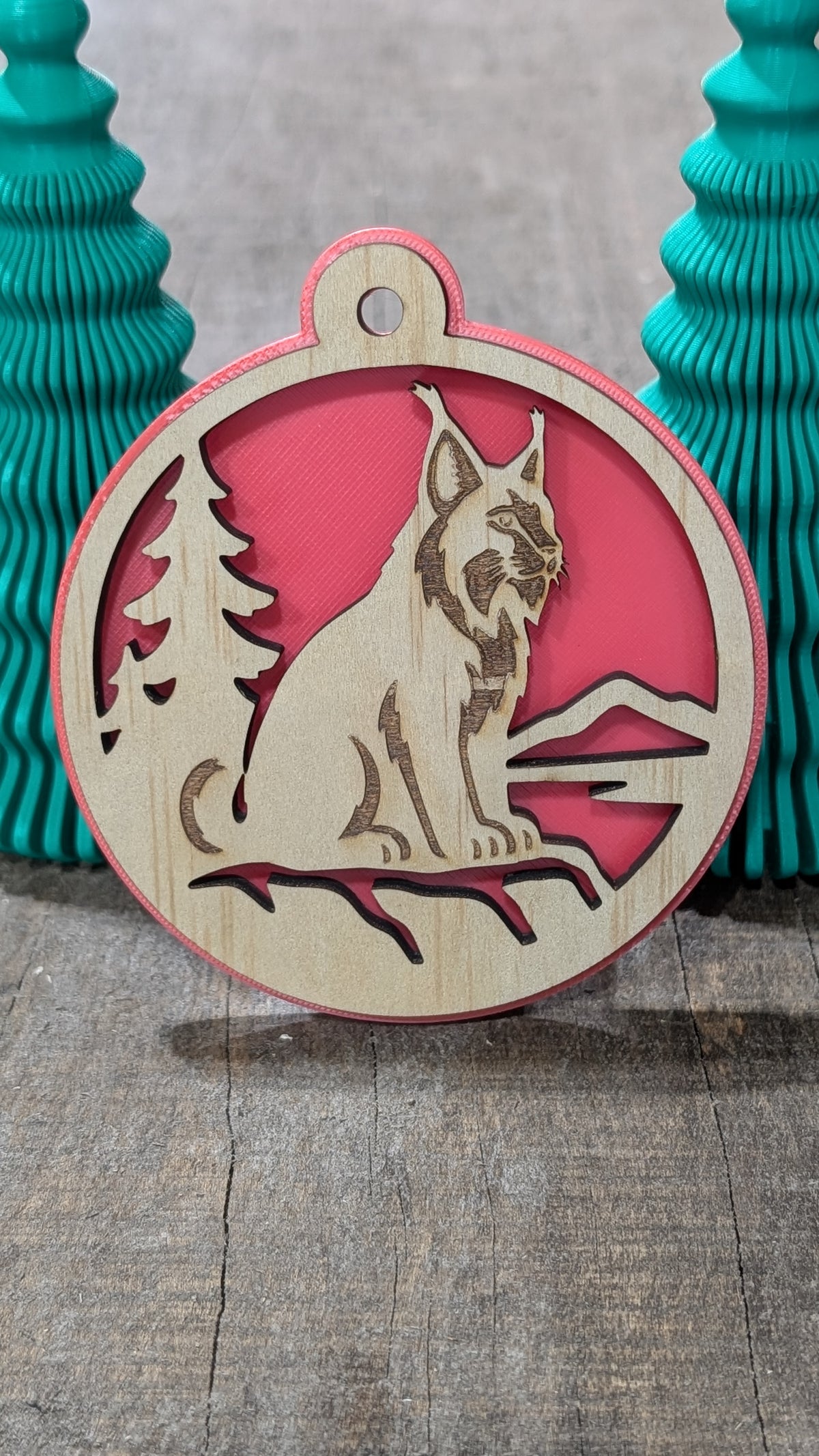 Boule de Noël en bois avec lynx, sapin et montagnes, sur fond rouge.