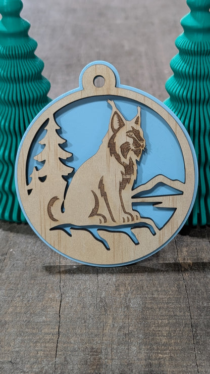 Boule de Noël en bois avec lynx, sapin et montagnes, sur fond bleu pâle.