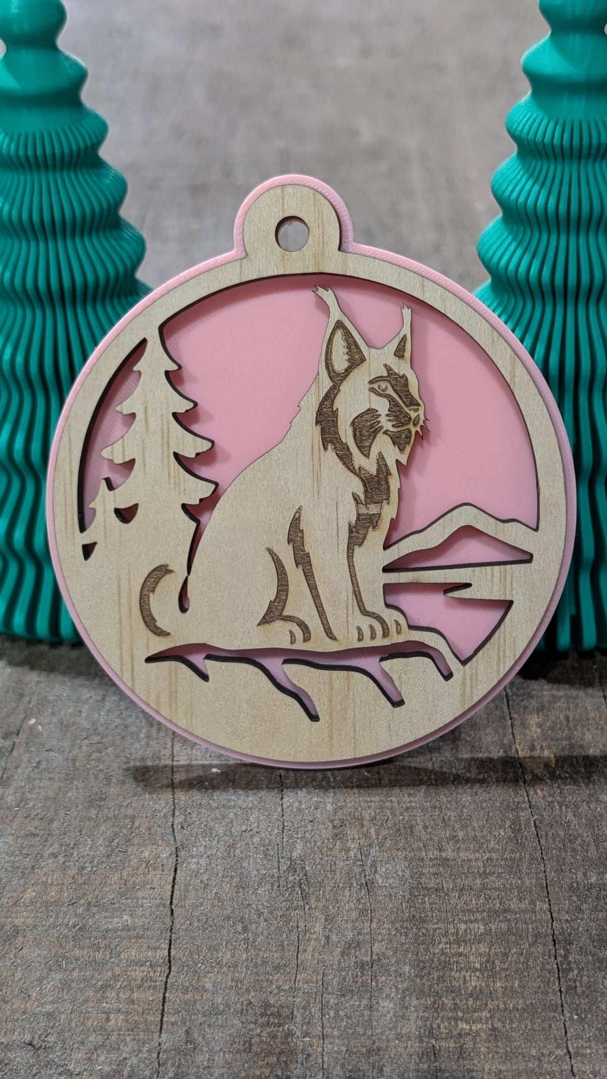 Boule de Noël en bois avec lynx, sapin et montagnes, sur fond rose.